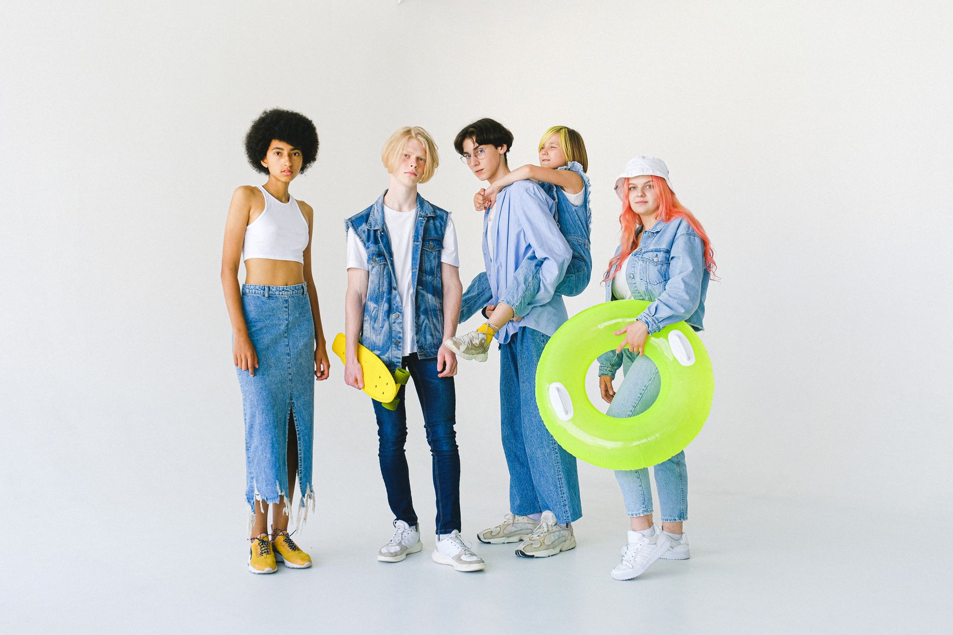 Full body of cool multiethnic teenagers and schoolchild in casual denim outfit standing in studio with inflatable ring and sk