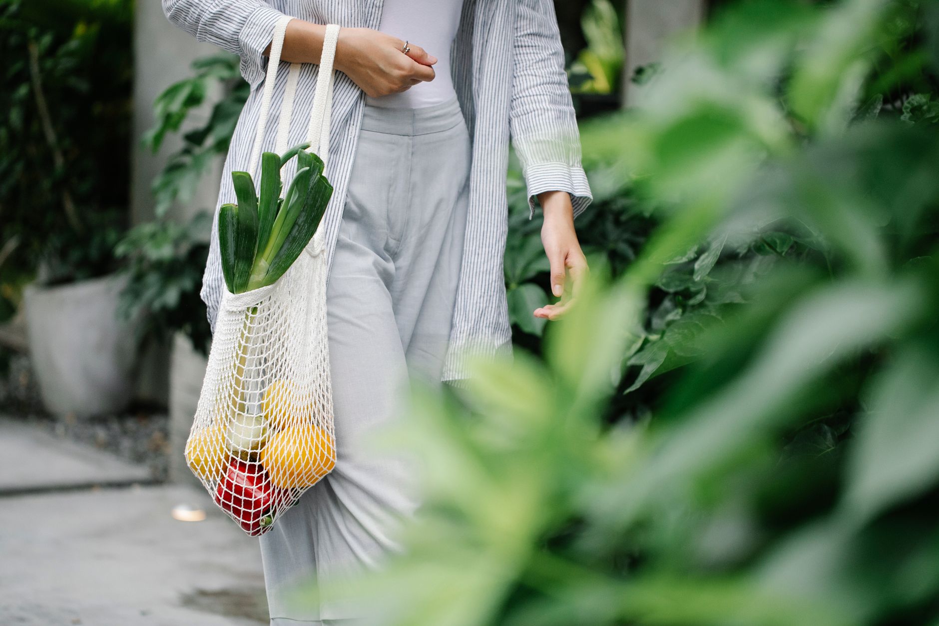 Crop anonymous female wearing casual outfit carrying net grocery bag with ripe vegetables including leek and capsicums in gar
